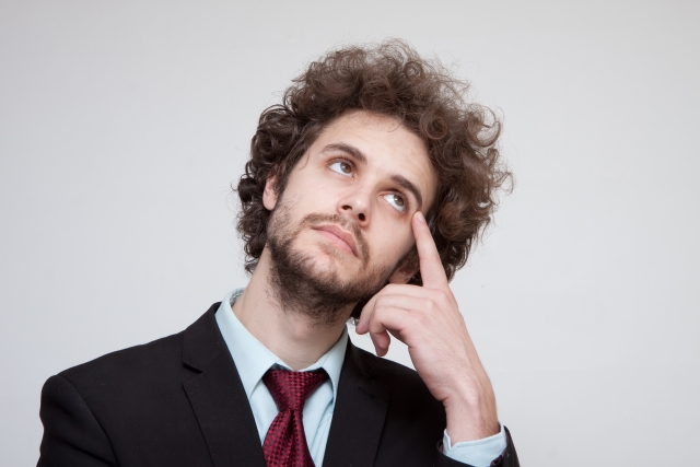 Man in black suit tilting his head to the right, with a pondering expression, and index finger resting at outer eye