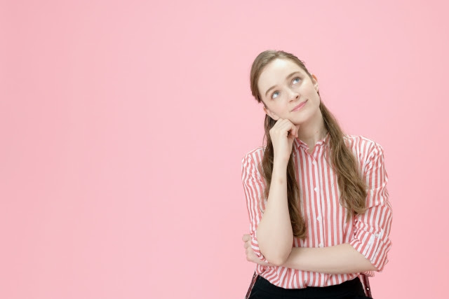 Woman with long brown hair and pink striped shirt in thinking pose, with pastel pink background
